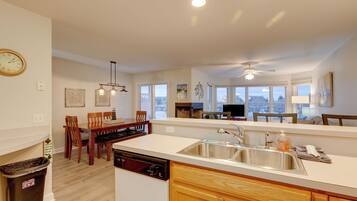 A modern kitchen with a dishwasher, sink, and wooden cabinets. There is a dining area with chairs and a table, and a view of the outside through large windows.