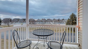 A covered balcony with two metal tables and chairs, overlooking a residential area with houses and a body of water.