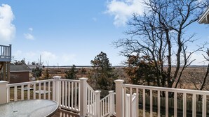 Una terraza de madera con una mesa de vidrio, barandillas blancas y vistas a un paisaje rural.