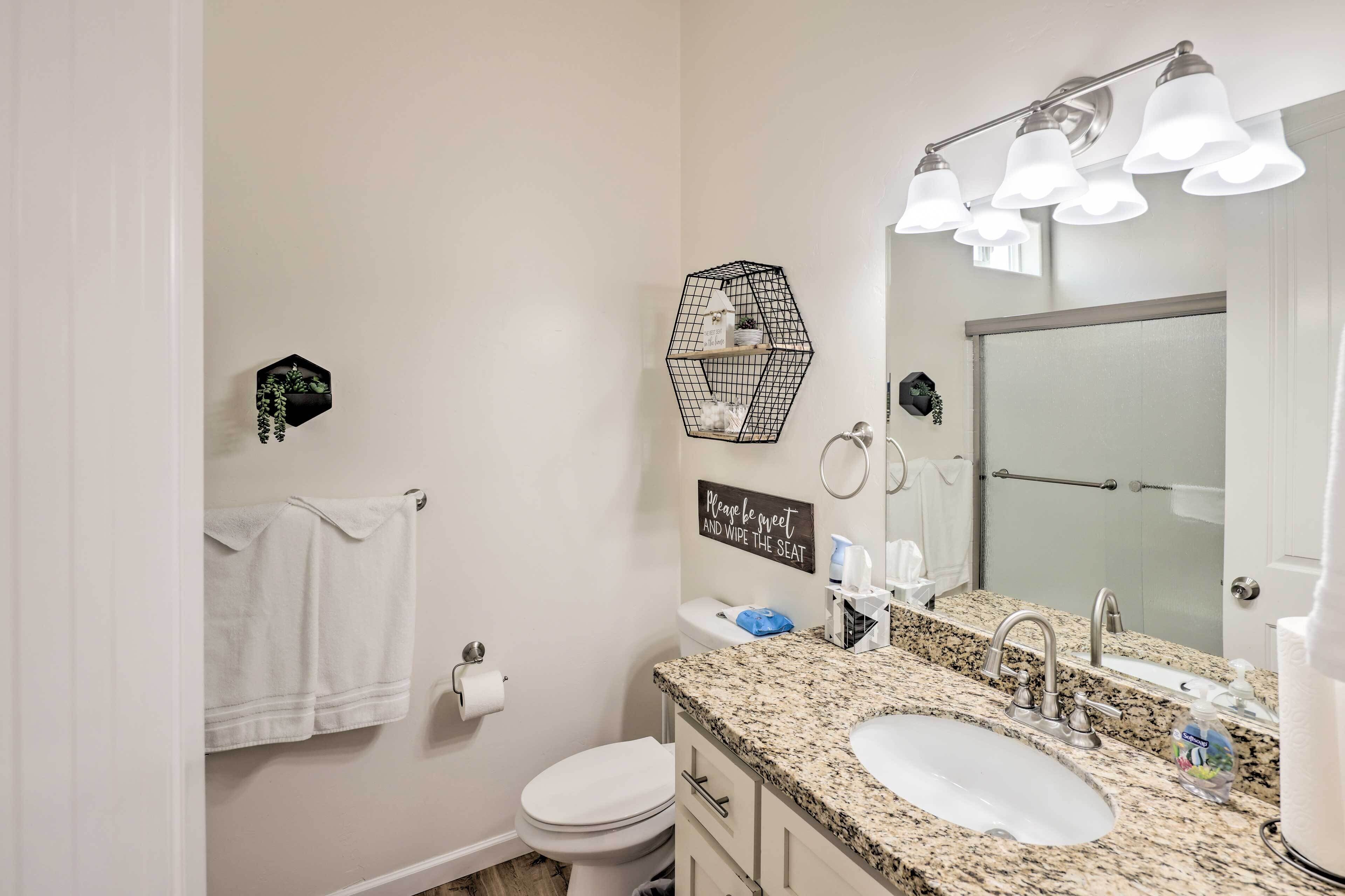 Une salle de bain avec des toilettes blanches, un lavabo surmonté d’un plan de travail en granit, un miroir avec éclairage intégré, un porte-serviettes et un élément décoratif hexagonal sur le mur.
