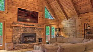 A log cabin interior with a stone fireplace, a large flat-screen TV mounted above it, a cozy seating area with a sofa, and a wooden ceiling. - 7 Mi to Ski Slopes: Mars Hill Cabin w/ Hot Tub! (Mars Hill)