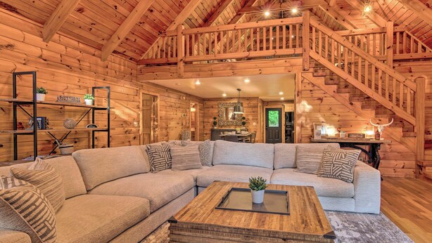 A log cabin interior with a large L-shaped sectional sofa, a wooden coffee table, and a staircase leading to an upper level. - 7 Mi to Ski Slopes: Mars Hill Cabin w/ Hot Tub! (Mars Hill)