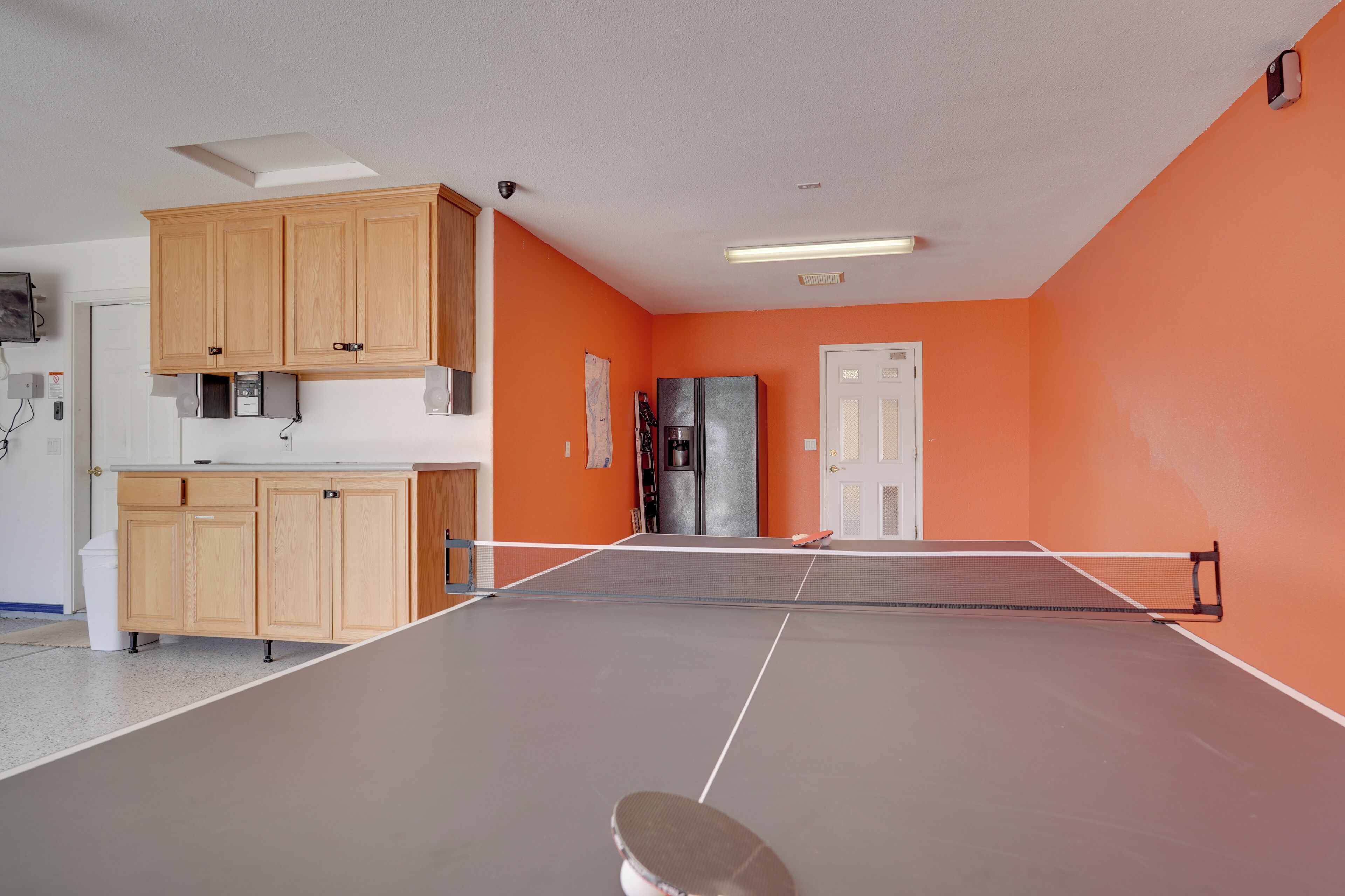 A room with a ping pong table, wooden cabinets, a refrigerator, and an orange wall.