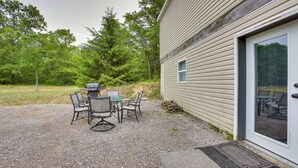 A gravel patio with a grill and chairs, a door with a glass panel, and a forested area in the background. - Spacious Wellston Home Near Hiking & Skiing! (Wellston)