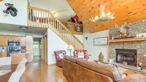 A spacious living area with a wooden staircase, a fireplace, and a kitchen in the background. - Game Room & Fire Pit: Picturesque Cabin in Murphy (Murphy)
