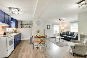 A modern kitchen with blue cabinets, a dining table with chairs, a living area with a sofa and armchair, and a clock on the wall. - Chic & Central Apartment ~ 6 Mi to Sebec Lake! (Dover-Foxcroft)