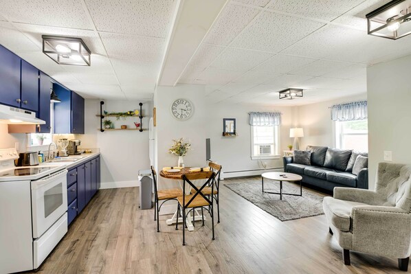 A modern kitchen with blue cabinets, a dining table with chairs, a living room with a sofa and armchair, and a clock on the wall. - Chic & Central Apartment ~ 6 Mi to Sebec Lake! (Dover-Foxcroft)