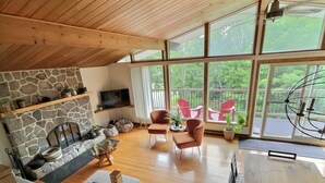 A spacious living room with a stone fireplace, wooden ceiling, and large windows offering a view of outdoor chairs.
