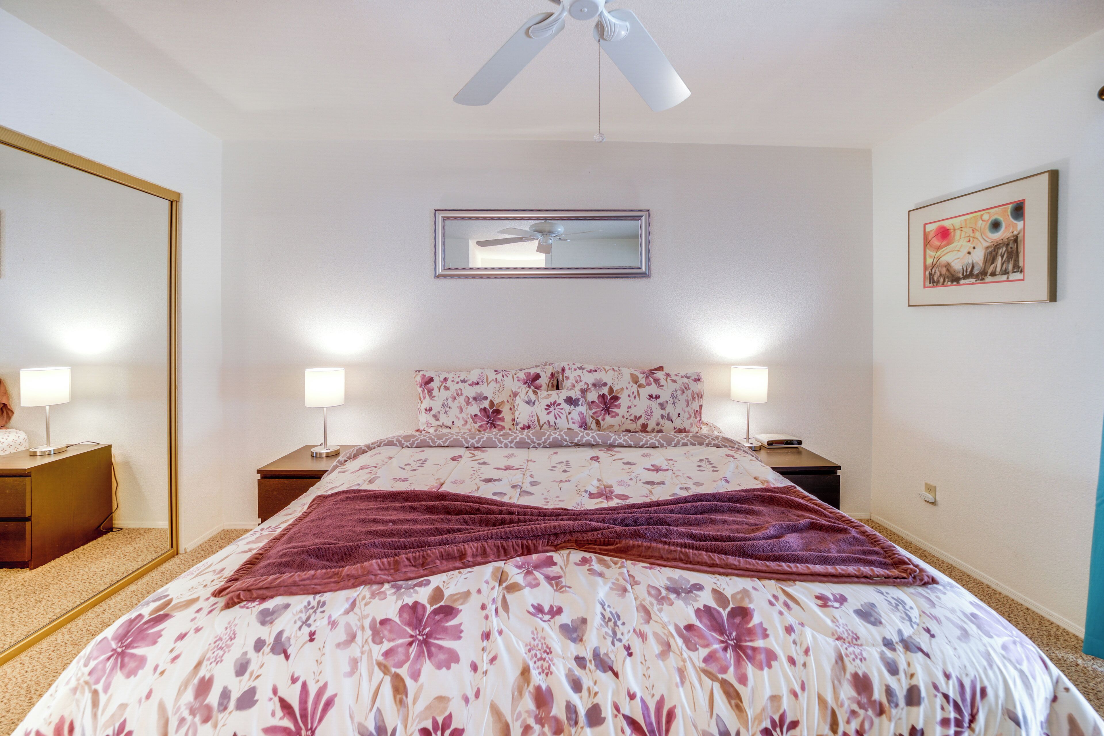 A bedroom with a bed, two bedside tables, a mirror, a ceiling fan, and a framed picture on the wall.