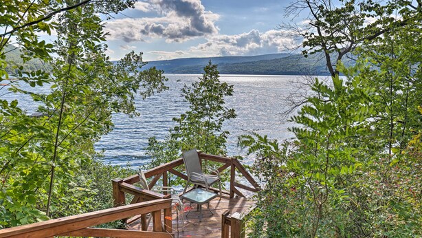 Une terrasse en bois avec une table et des chaises en verre, donnant sur un lac et entourée de verdure.