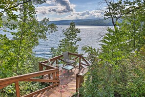 Um deck de madeira com mesa e cadeiras de vidro, com vista para um lago, rodeado por vegetação.