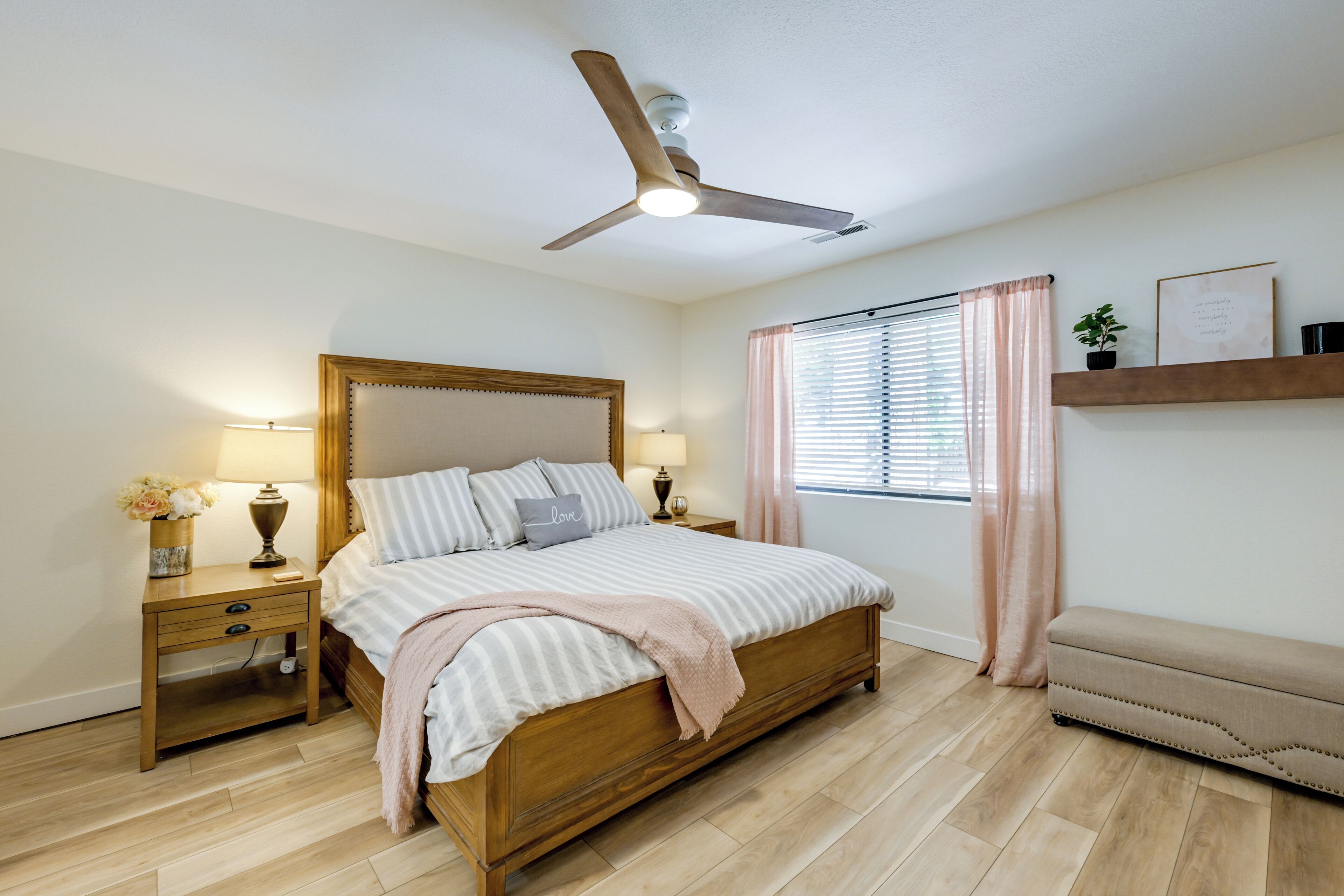 A bedroom with a wooden bed, bedside tables, a ceiling fan, a window with curtains, and a shelf with a plant.