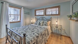 A bedroom with a bed, two windows with blinds, a bedside table with a lamp, and a wall-mounted shelf with plants. - Locust Lake Village Cabin w/ Deck & Lake View! (Pocono Lake)