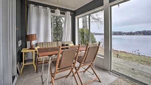 A dining area with a wooden table and chairs, a lamp, and a view of a lake through large windows. - Waterfront Cable Lake Cabin w/ Grill! (Dowagiac)