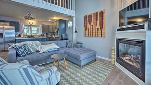 A spacious living room with a grey sectional sofa, a fireplace, and a kitchen in the background. - Waterfront Cable Lake Cabin w/ Grill! (Dowagiac)