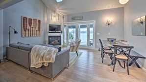 A spacious living room with a gray sofa, a fireplace, a dining area with a table and chairs, and a large window overlooking a scenic view. - Waterfront Cable Lake Cabin w/ Grill! (Dowagiac)