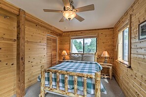 A wooden cabin bedroom with a bed, two bedside lamps, a ceiling fan, and a window with a view of trees. - Overgaard Cabin w/ Hot Tub, Fire Pit & Deck! (Overgaard)