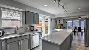 Una cocina moderna con armarios grises, un lavamanos de acero inoxidable y una isla grande. Se ve un comedor con mesa y sillas a través de la ventana.