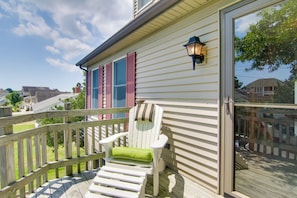 A wooden deck with a white chair and a green cushion, a glass door, and a window with red shutters.