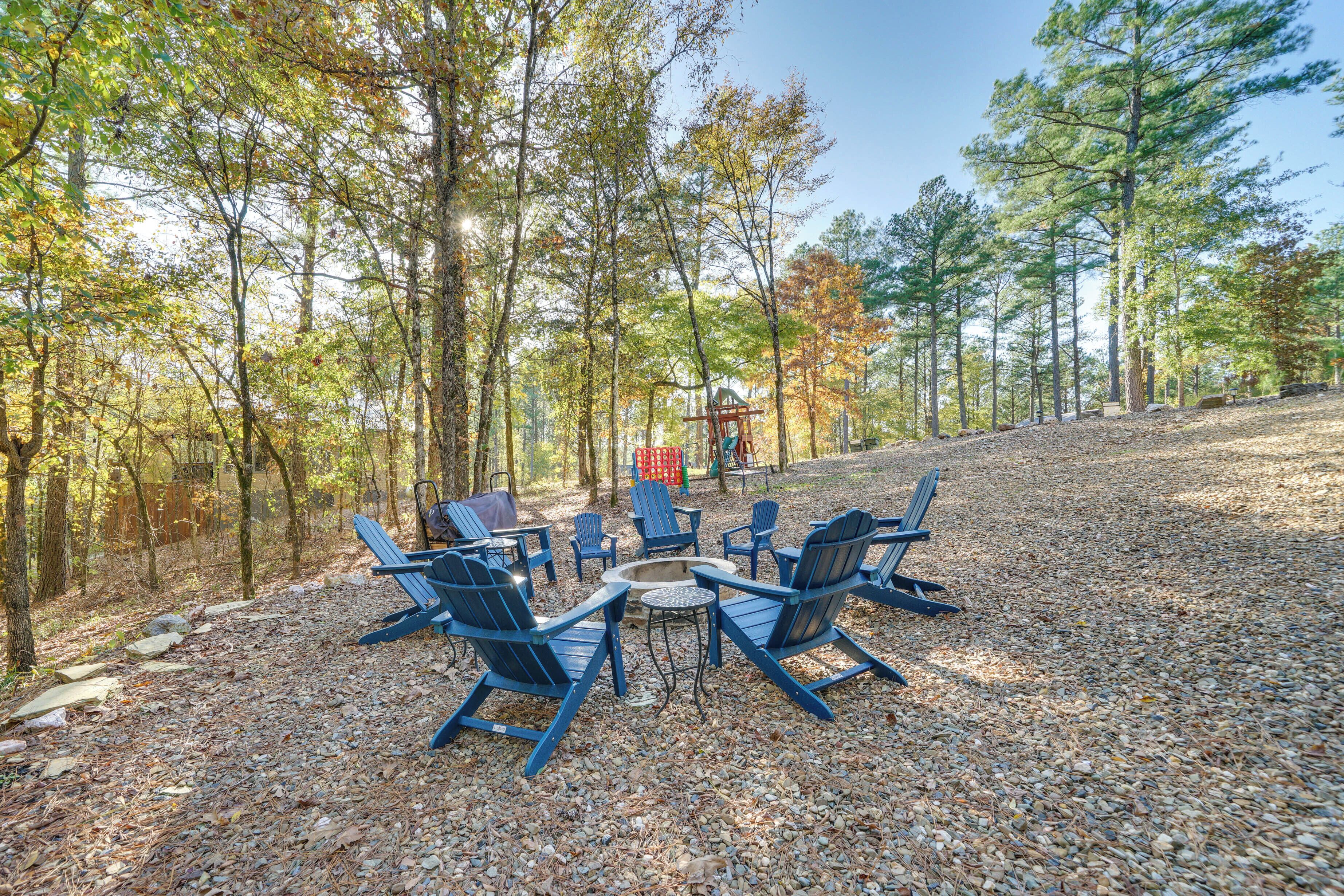 A circular seating arrangement with chairs around a central fire pit, set on a gravel area surrounded by trees.