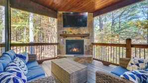 A covered deck with a stone fireplace, a flat-screen TV, wicker furniture, and a view of a forest. - Furnished Deck, Hot Tub! Family Gem in Broken Bow (Broken Bow)