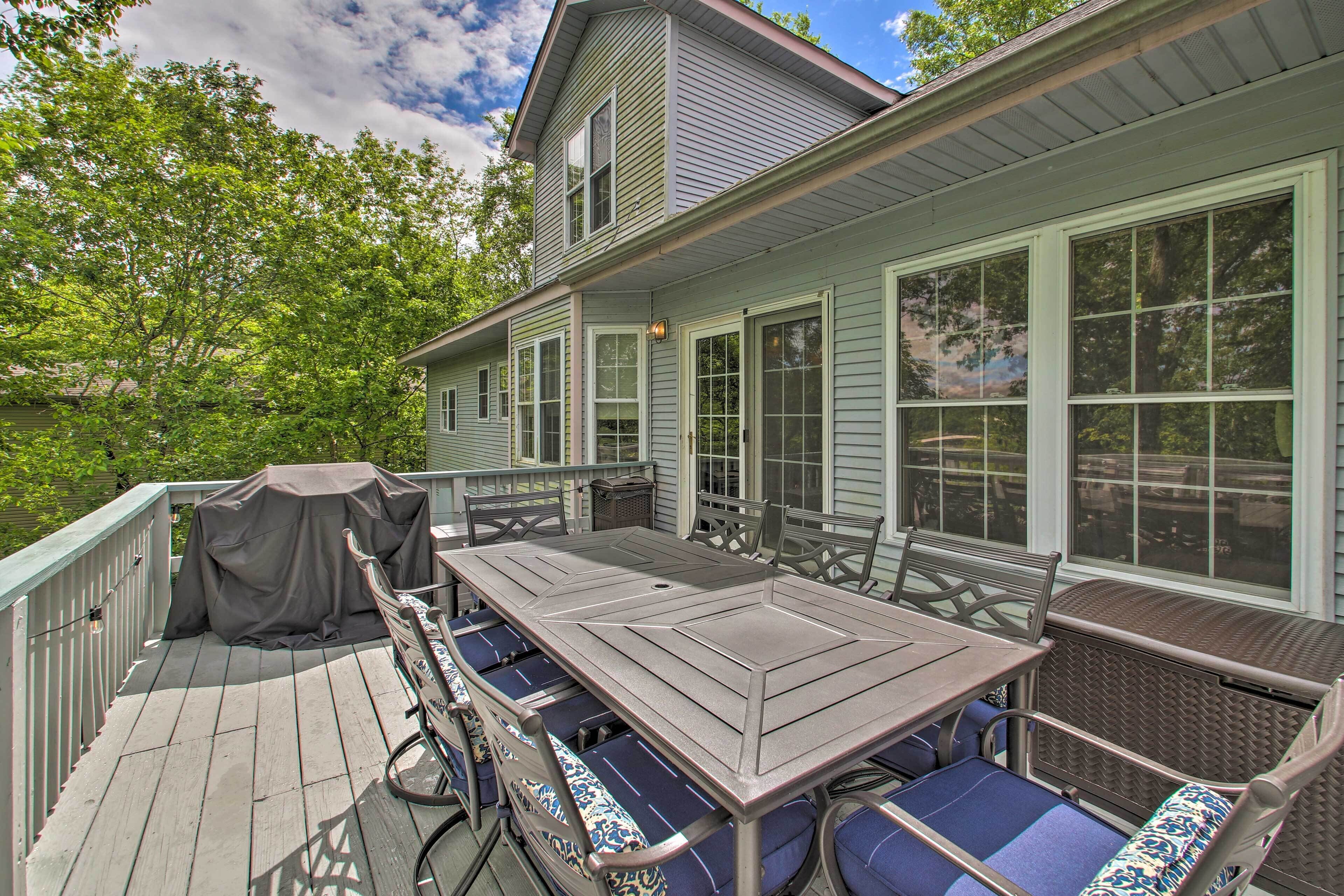 Une terrasse extérieure couverte, équipée d’une table et de chaises, d’un barbecue et offrant une vue sur une maison dotée de grandes fenêtres.