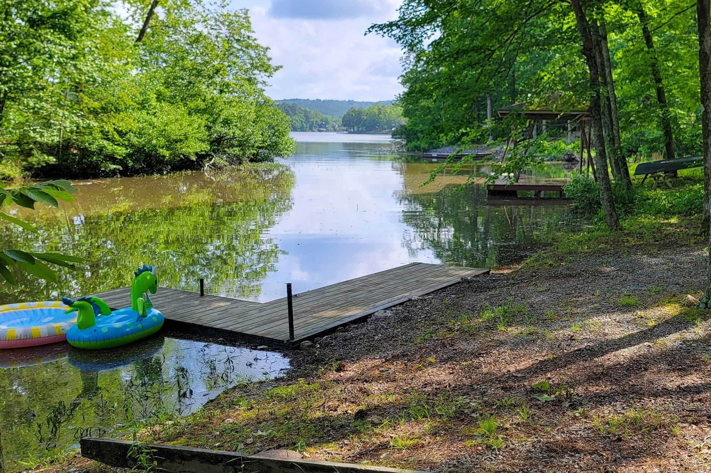 Un lac paisible, avec une jetée en bois qui s’avance sur l’eau, entouré d’une végétation luxuriante et d’arbres.