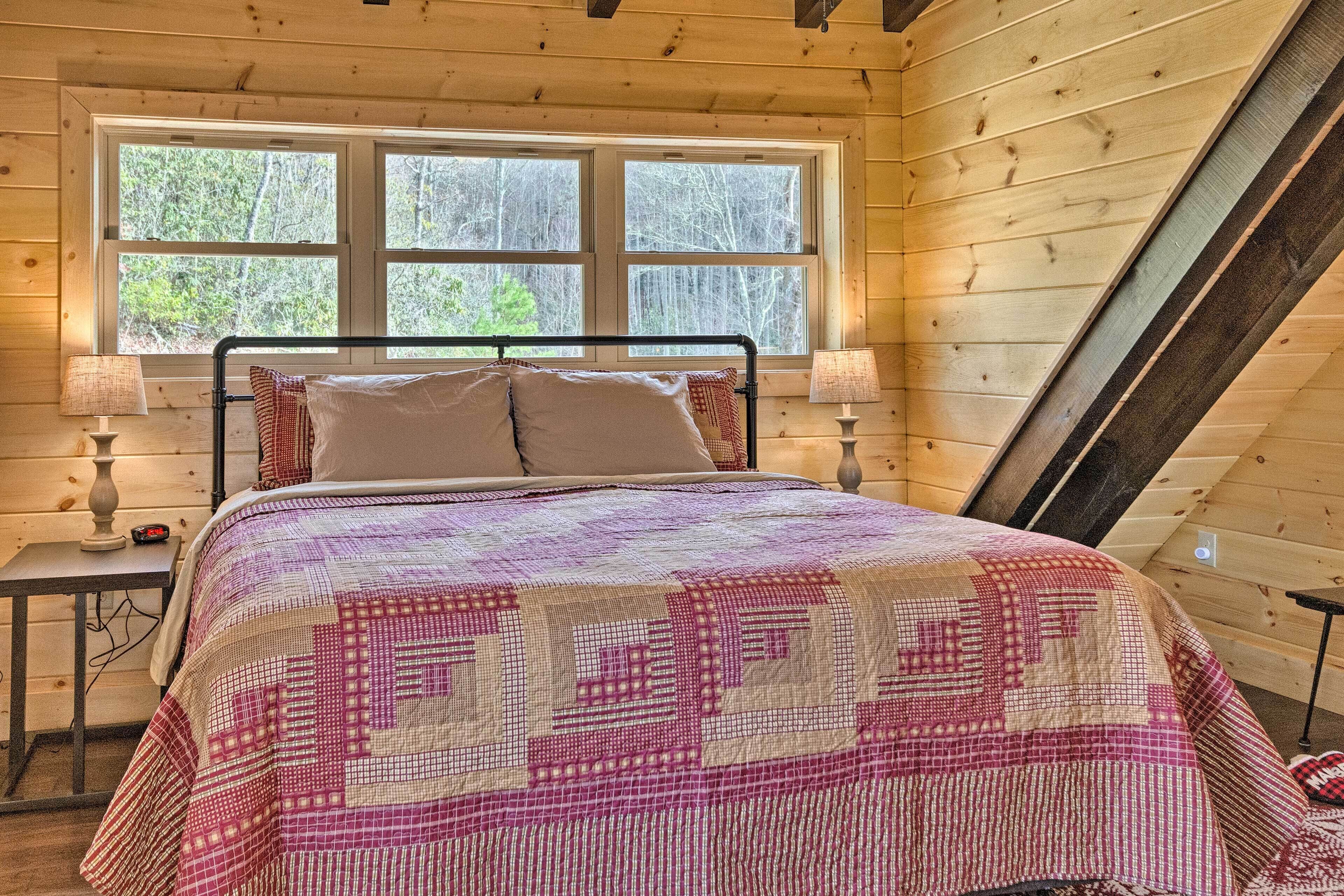 A wooden cabin bedroom with a bed, two bedside lamps, and a window with a view of trees.