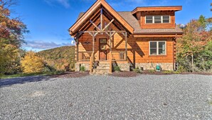 A two-story log cabin with a stone foundation, a front porch, and a clear view of the surrounding forest. - Hilltop Cabin on 5 Acres w/ Hot Tub & Waterfall! (Bryson City)