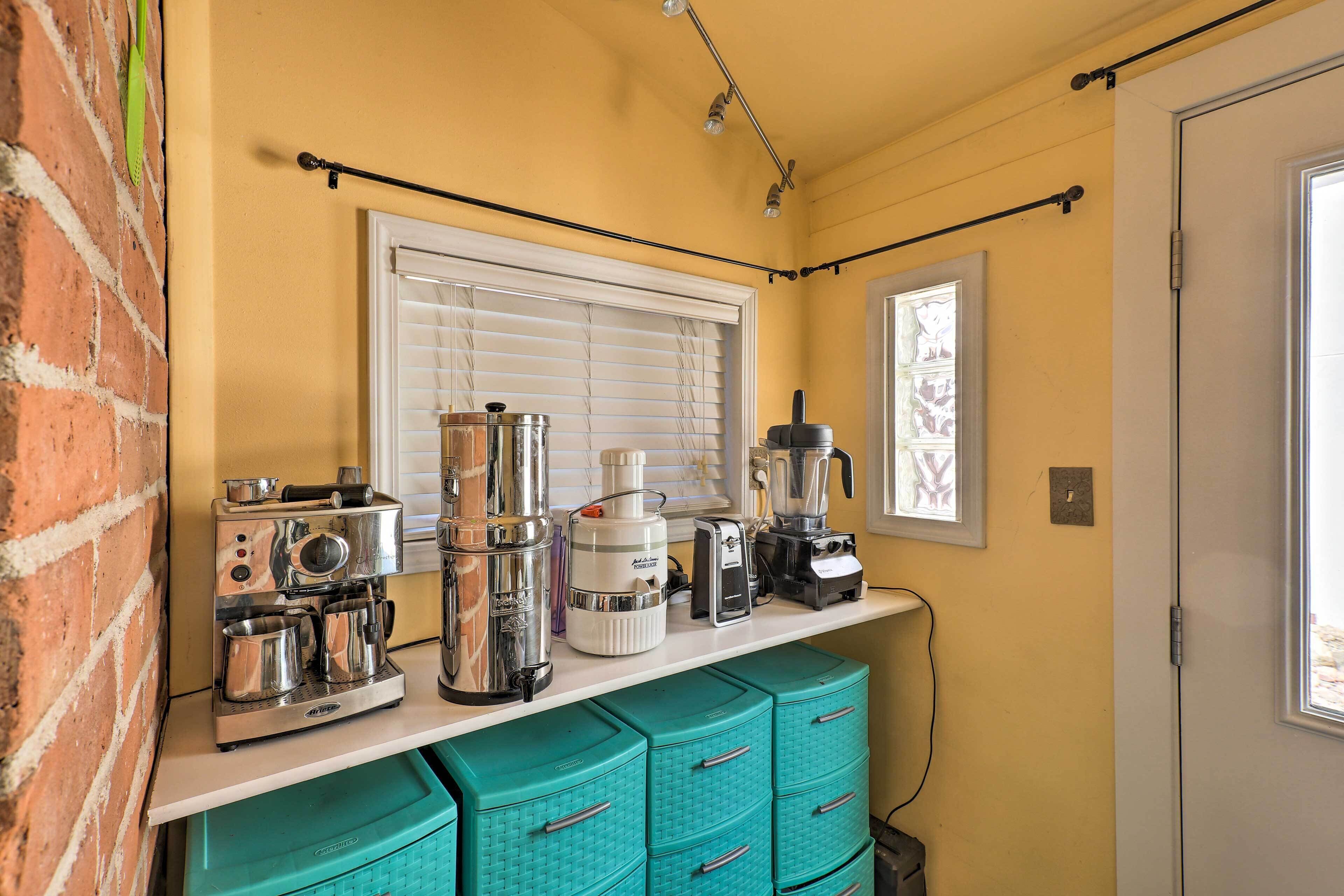 A compact kitchen with a coffee maker, blender, and toaster on a white countertop. There are teal drawers below and a window with blinds.