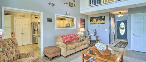 A spacious living room with a sofa, ottoman, and coffee table, a kitchen in the background, and a balcony with railings.