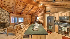 A log cabin interior with a stone fireplace, wooden furniture, a kitchen with stainless steel appliances, and a large window. - 11 Mi to Rainbow Lake: Family Home w/ Large Yard (Pinetop)