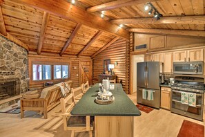 A log cabin interior with a stone fireplace, wooden furniture, a kitchen with stainless steel appliances, and a large window. - 11 Mi to Rainbow Lake: Family Home w/ Large Yard (Pinetop)