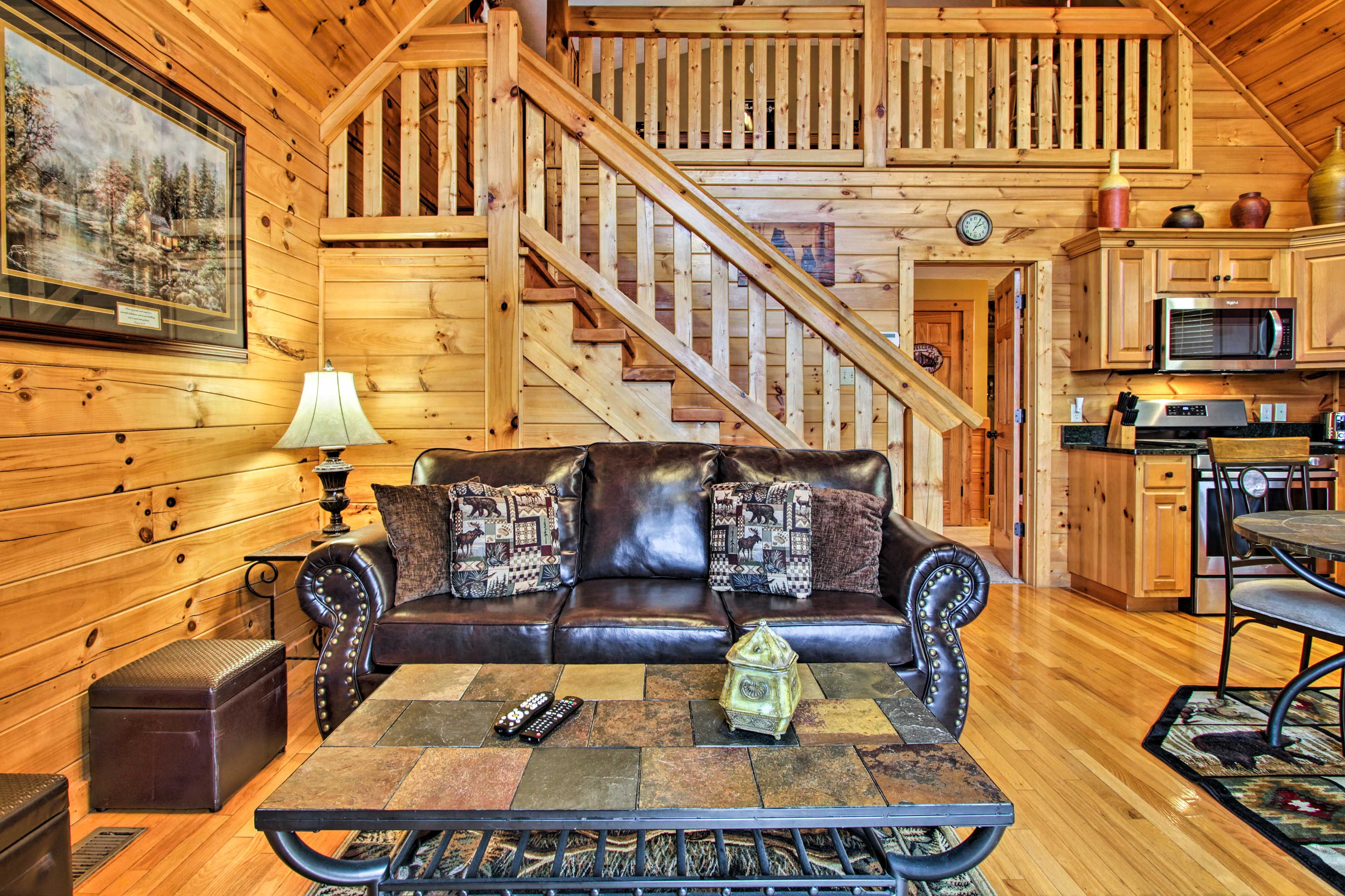 A wooden cabin interior with a leather sofa, a coffee table, a staircase, and a kitchen area.