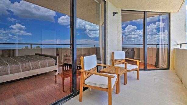 A balcony with a bed, two wooden chairs, and a view of the sky and clouds through large glass doors. - Modern Waterfront Hudson Condo w/ Resort Pool! (Hudson)