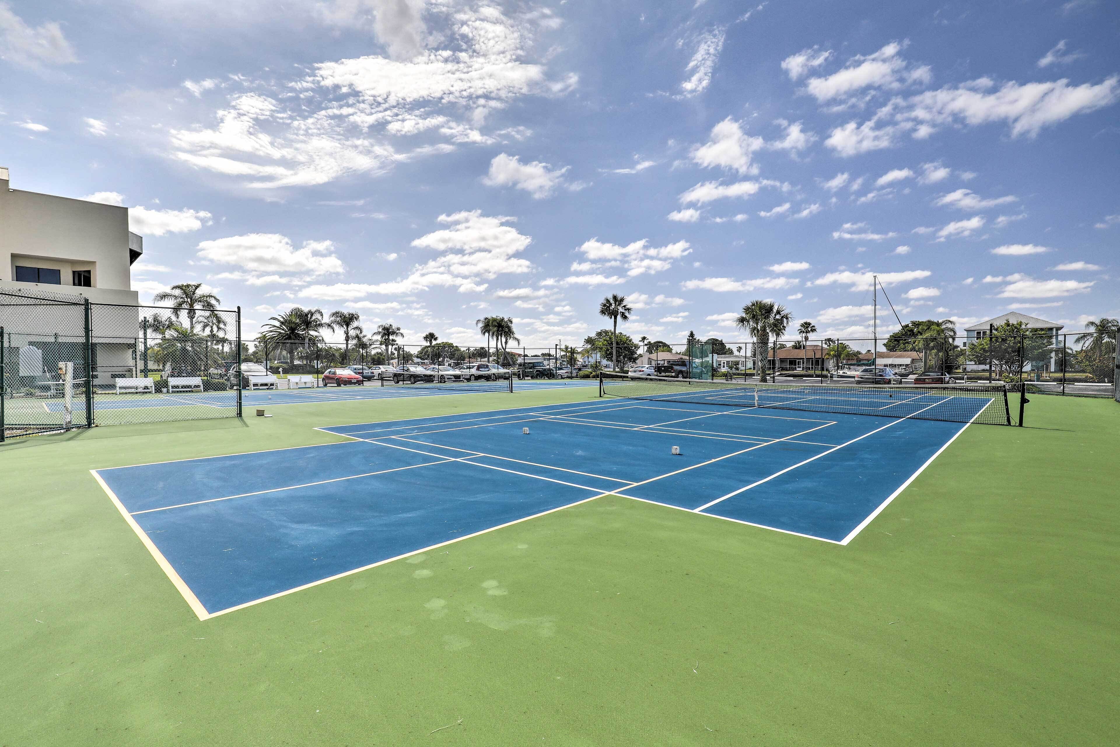 Una cancha de tenis con una red, rodeada por una cerca, palmeras y edificios al fondo.