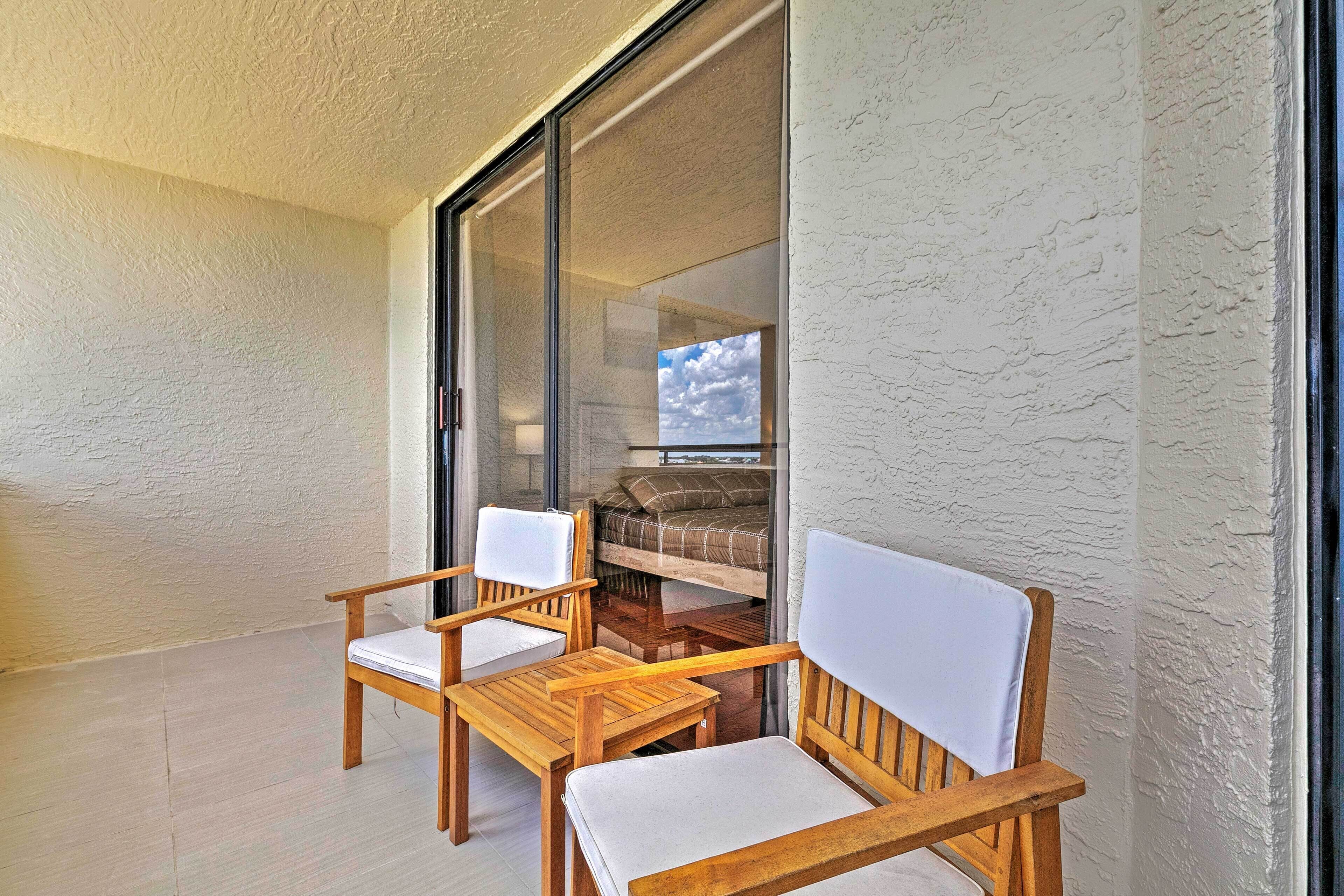 Un balcón con dos sillas de madera y una mesa, una puerta corrediza de vidrio que da a un dormitorio con cama y ventana.