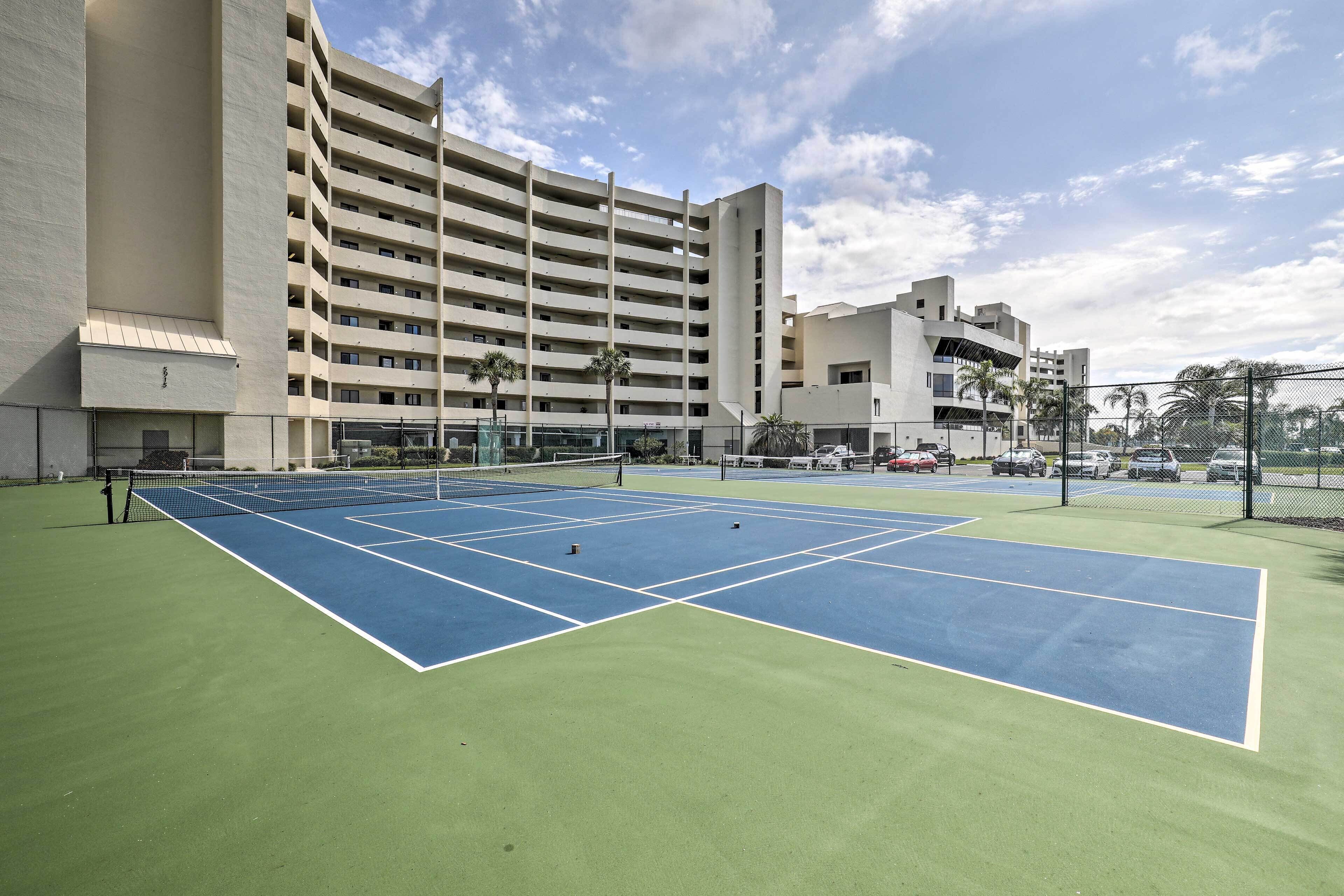 A tennis court with a net, surrounded by a fence, and adjacent to a multi-story building.