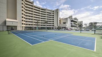 A tennis court with a net, surrounded by a fence, and adjacent to a multi-story building.
