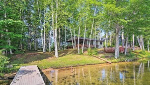 A wooden dock leading to a house surrounded by trees and a pond. - Lakefront Cabin w/ Private Dock & Beach! (Manistique)