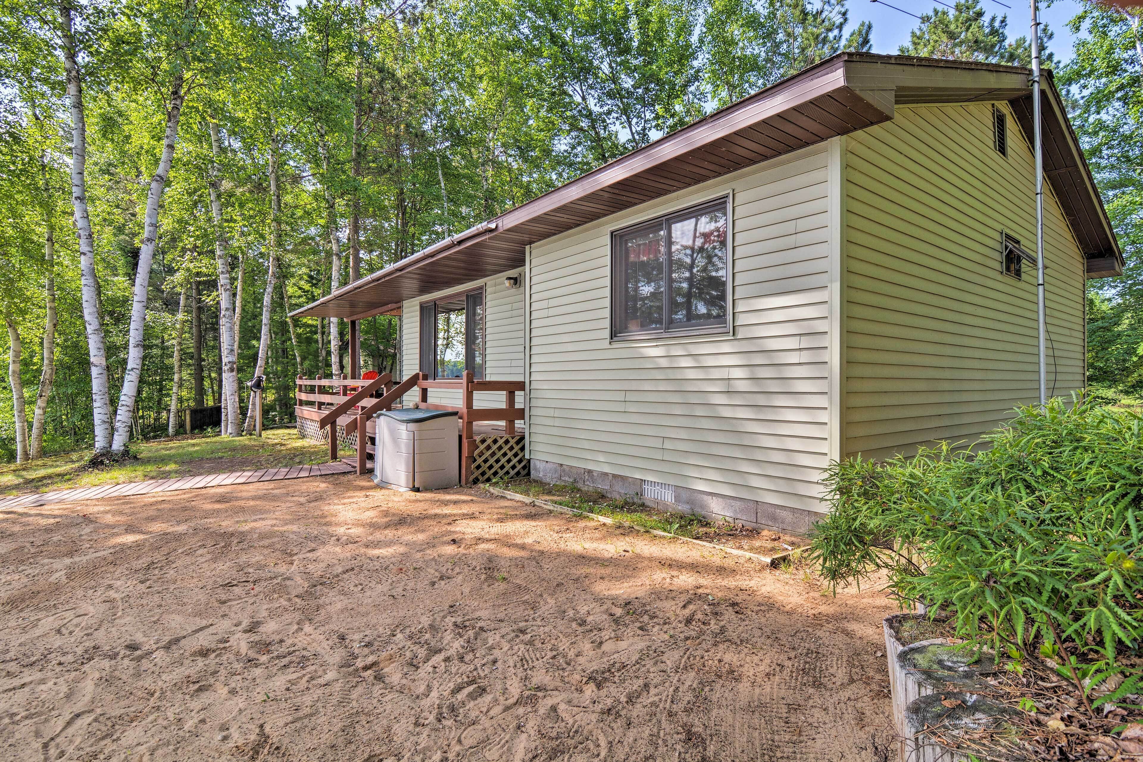 A small, single-story cabin with a wooden deck and a covered porch, surrounded by trees.