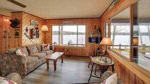 A cozy cabin interior with a wooden interior, a sofa, a table, and a large window overlooking a body of water. - Lakefront Cabin w/ Private Dock & Beach! (Manistique)