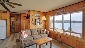 A cabin with a wooden interior, a sofa, a table, and a view of a lake. - Lakefront Cabin w/ Private Dock & Beach! (Manistique)