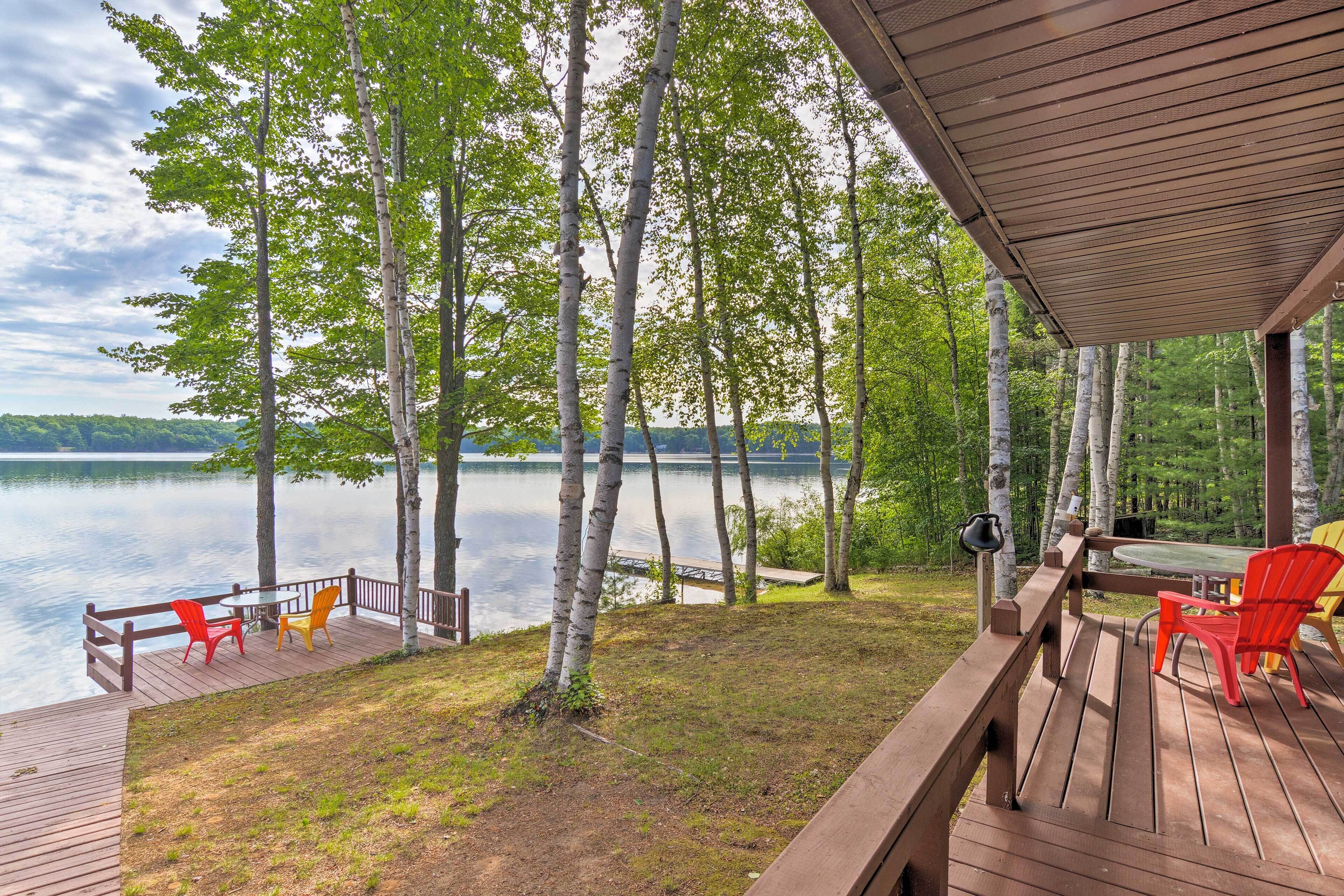 A wooden deck with outdoor seating, overlooking a calm lake surrounded by birch trees.