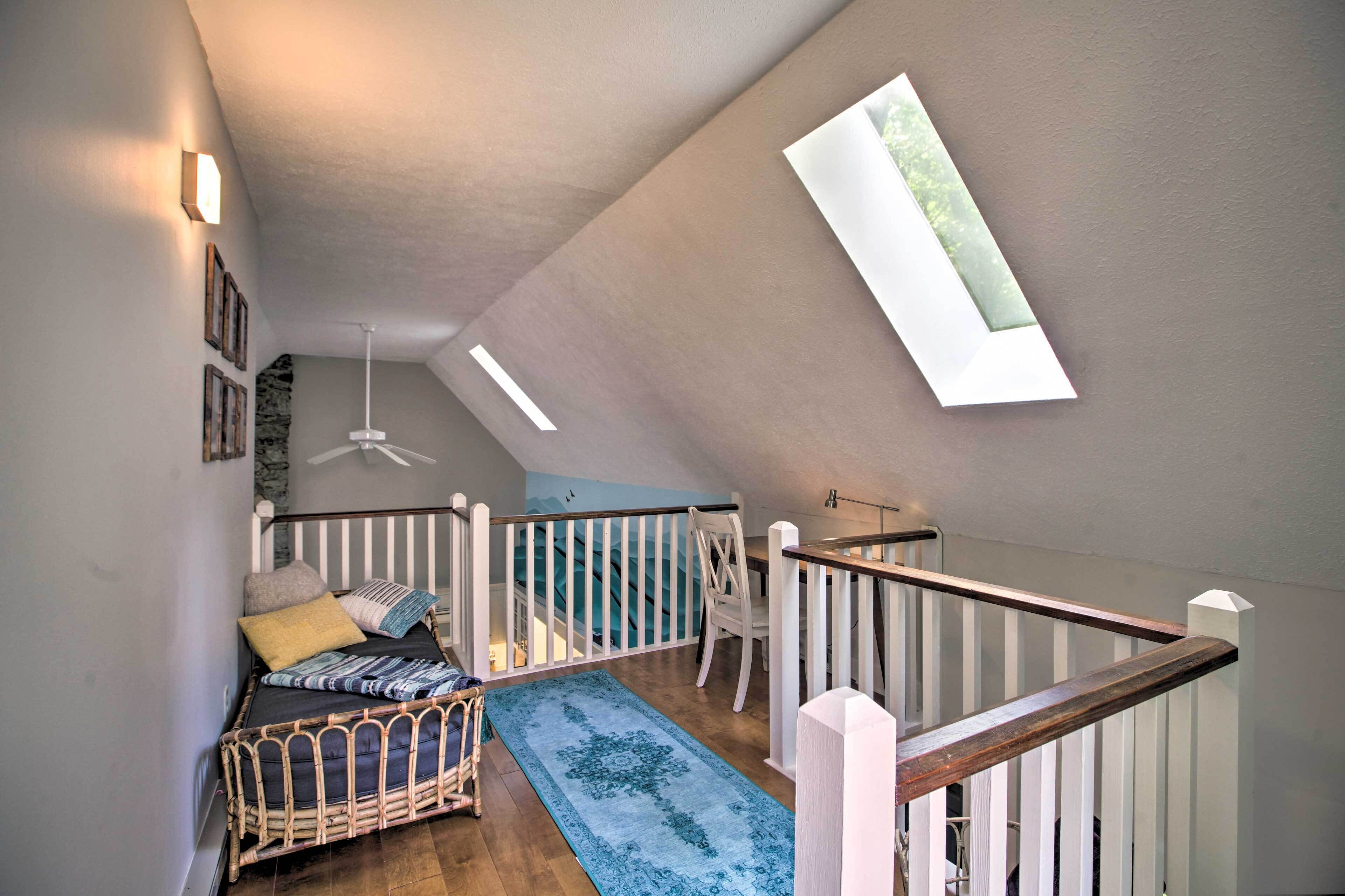 Dormitorio estilo loft con techo inclinado, una cama, una silla, barandal de madera y tragaluz.
