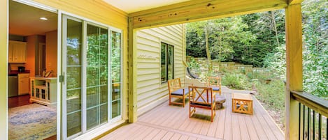 A wooden deck with a sliding glass door leading to a kitchen, a dining area with chairs and a table, and a view of a wooded area.