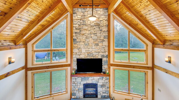 A room with a stone fireplace, wooden beams, and large windows offering a view of the outdoors. - Spacious & Secluded Forksville Home: Fire Pit (Forksville)