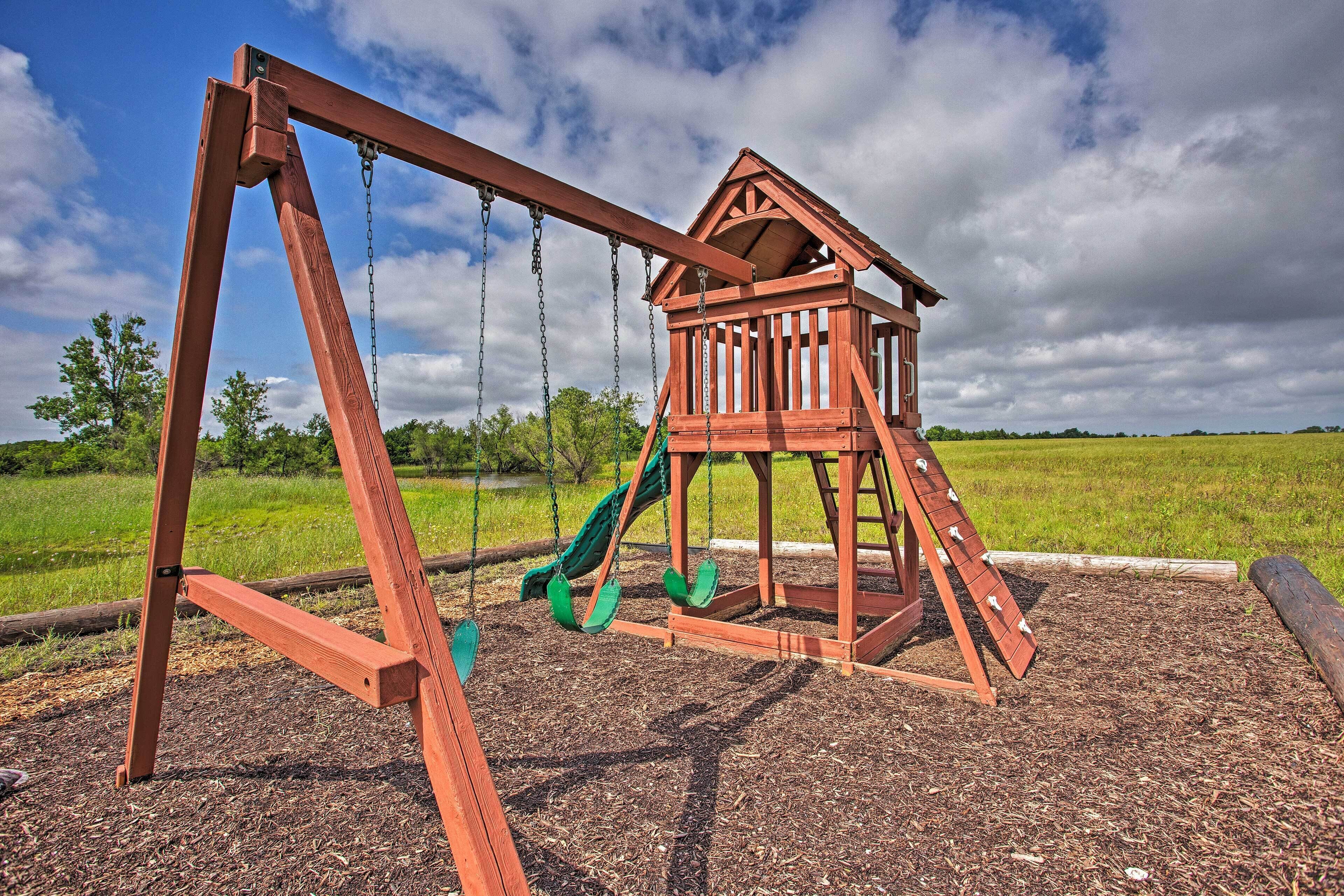 A wooden swing set with a slide, set on a mulch-covered ground, surrounded by a grassy field and trees under a partly cloudy sky.