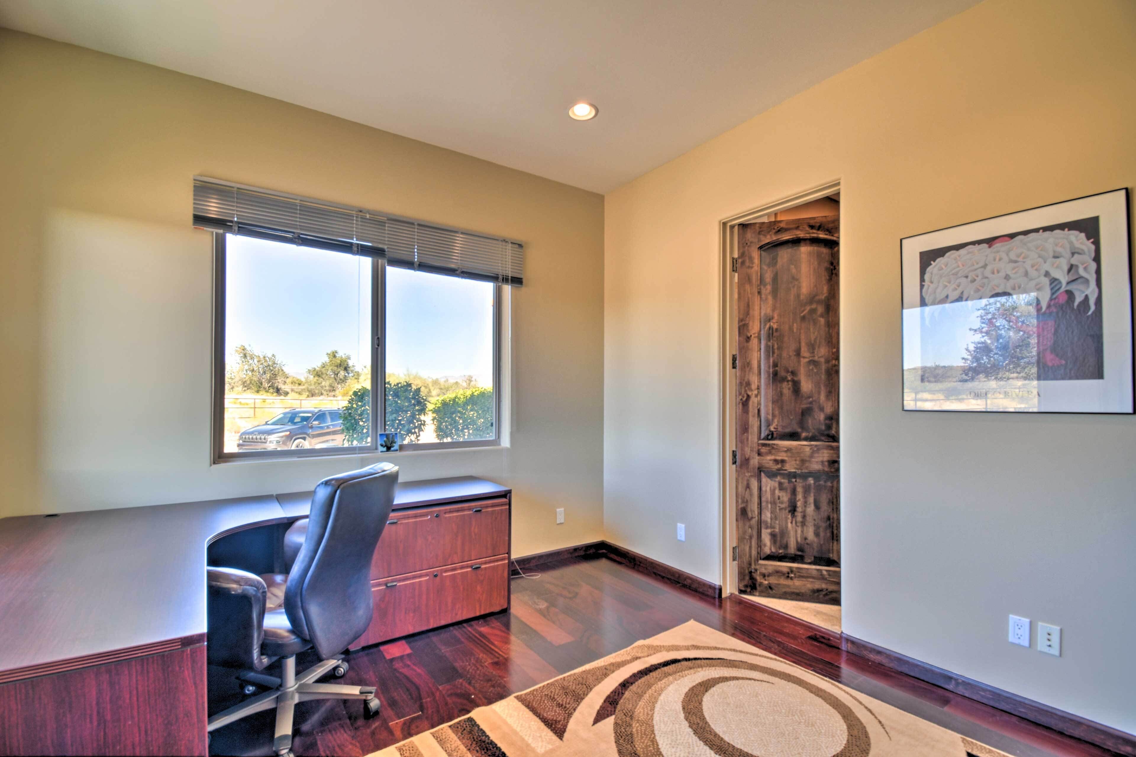A room with a desk, chair, large window, wooden door, and a framed picture on the wall.