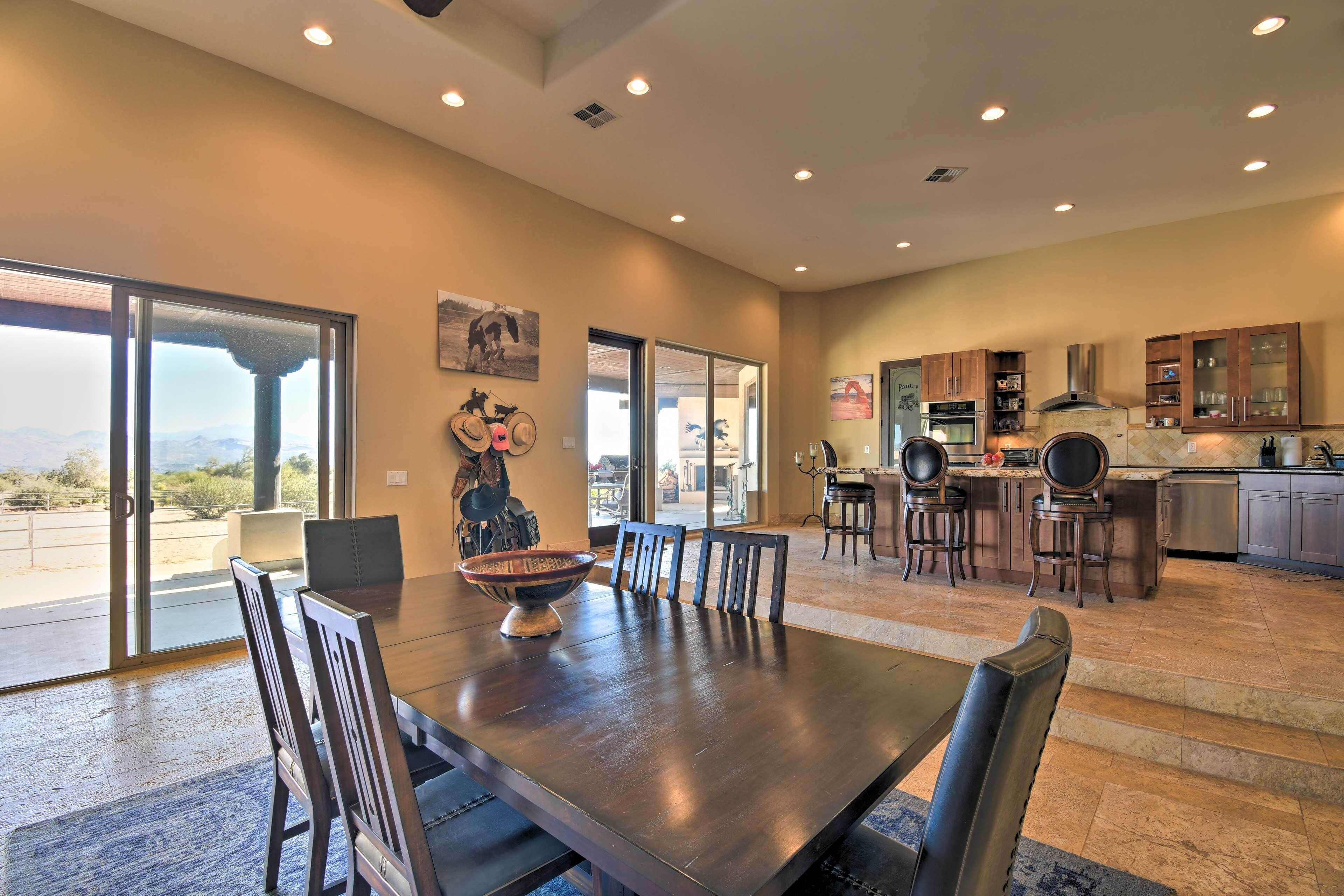 A spacious dining area with a large table and chairs, a kitchen in the background, and a view through a glass door.